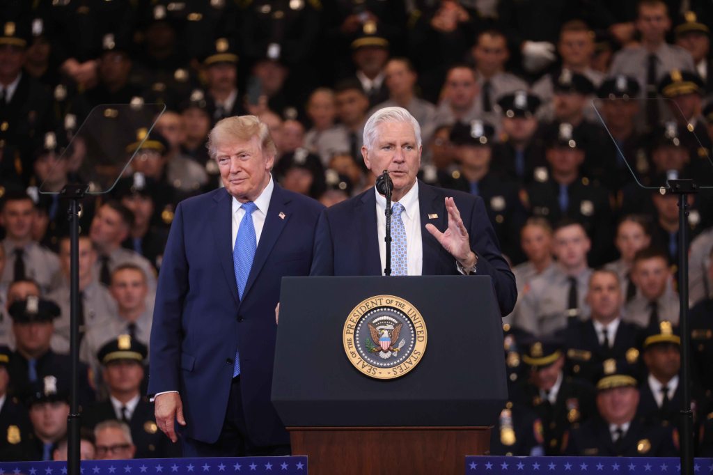 Bruce Blakeman speaks at a podium with Donald Trump standing beside him, in front of a crowd of police officers.