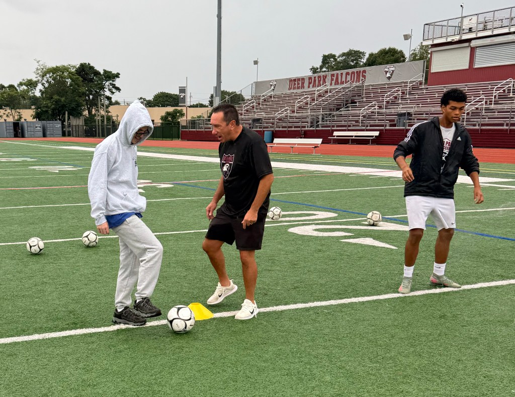 Coach Frank Vertullo stands on a soccer field, instructing a young player in a hoodie while another player looks on nearby.