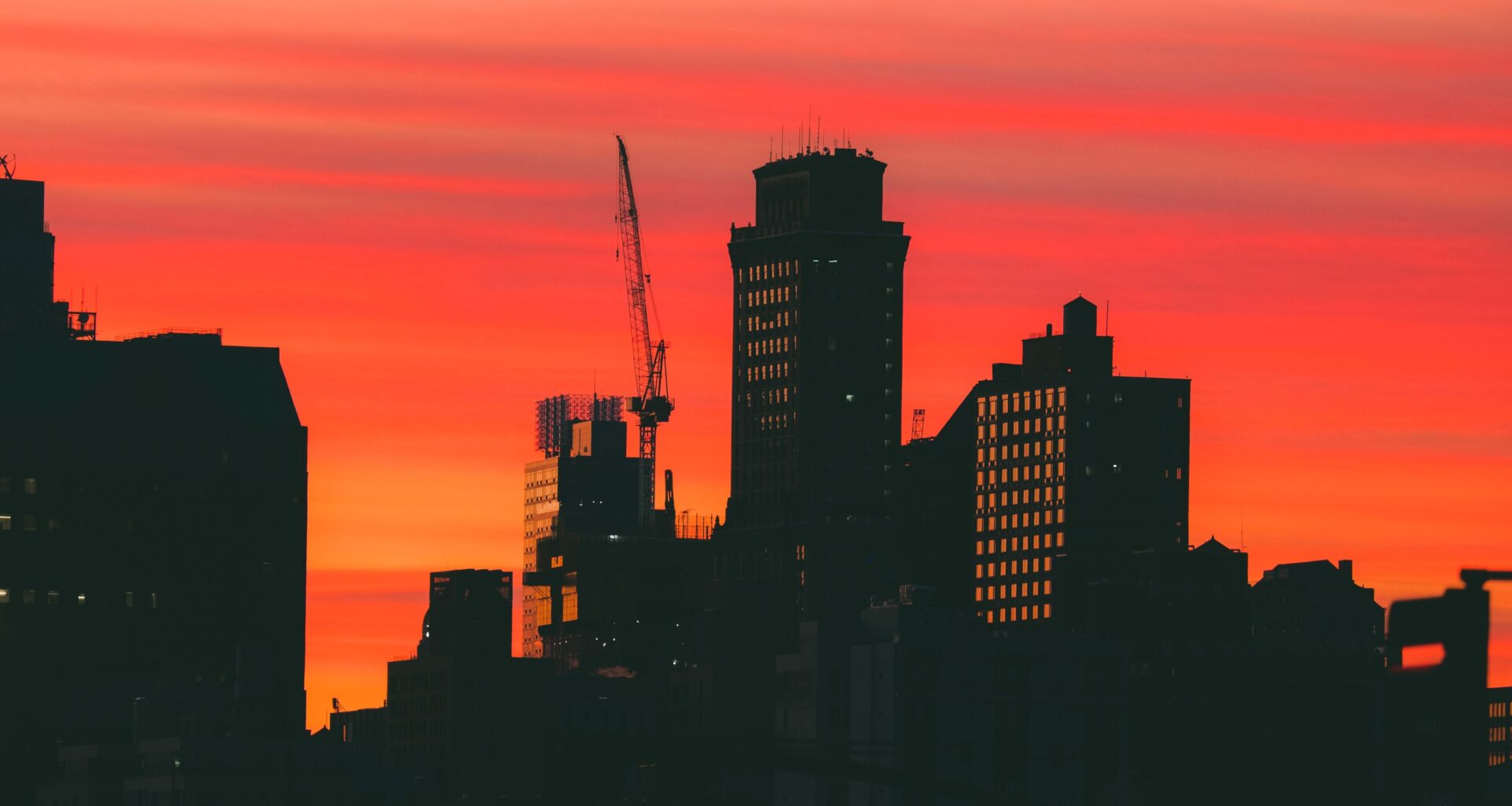 Silhouette of city buildings and a crane against a vivid orange and red sunset sky.