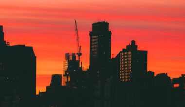 Silhouette of city buildings and a crane against a vivid orange and red sunset sky.