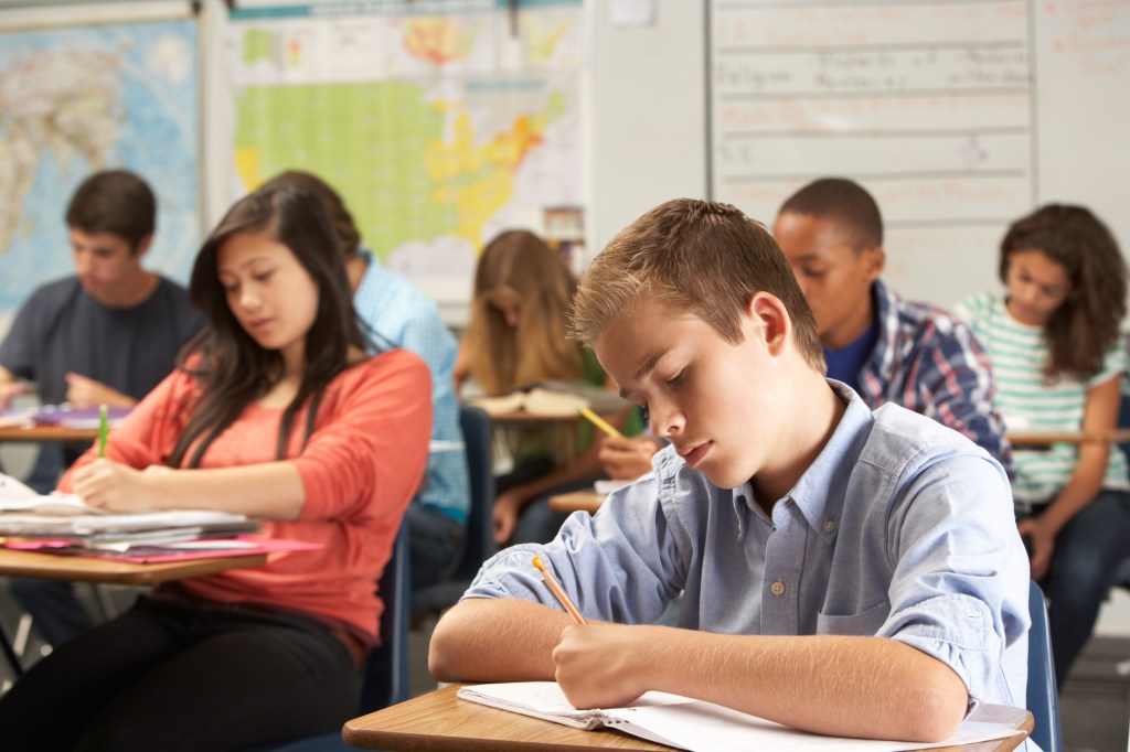 A young boy in a blue shirt sits at his desk, focused on writing in a notebook, while other high school students study in the background of a bright classroom.