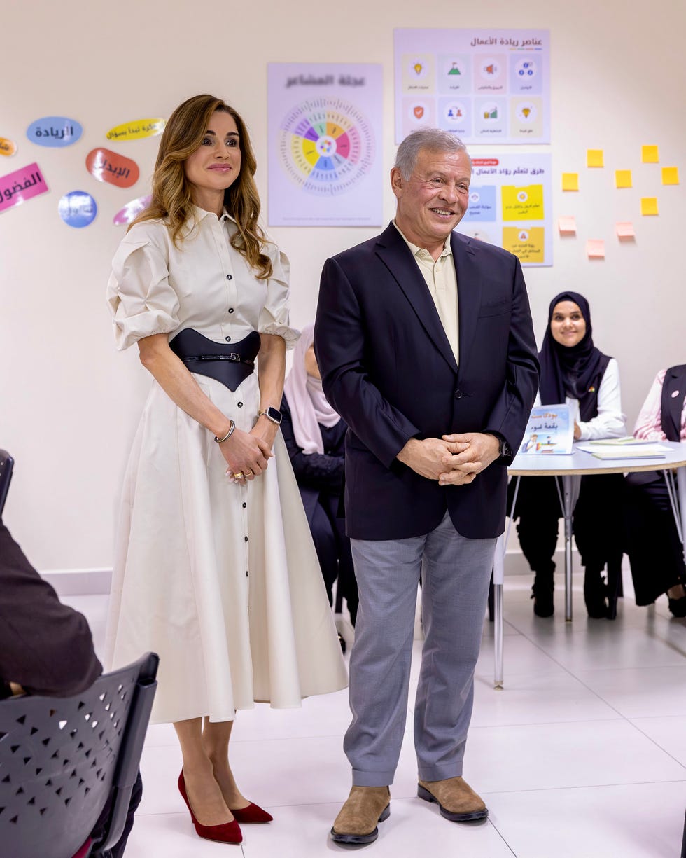 Two people standing together in a classroom setting with students and educational materials in the background.