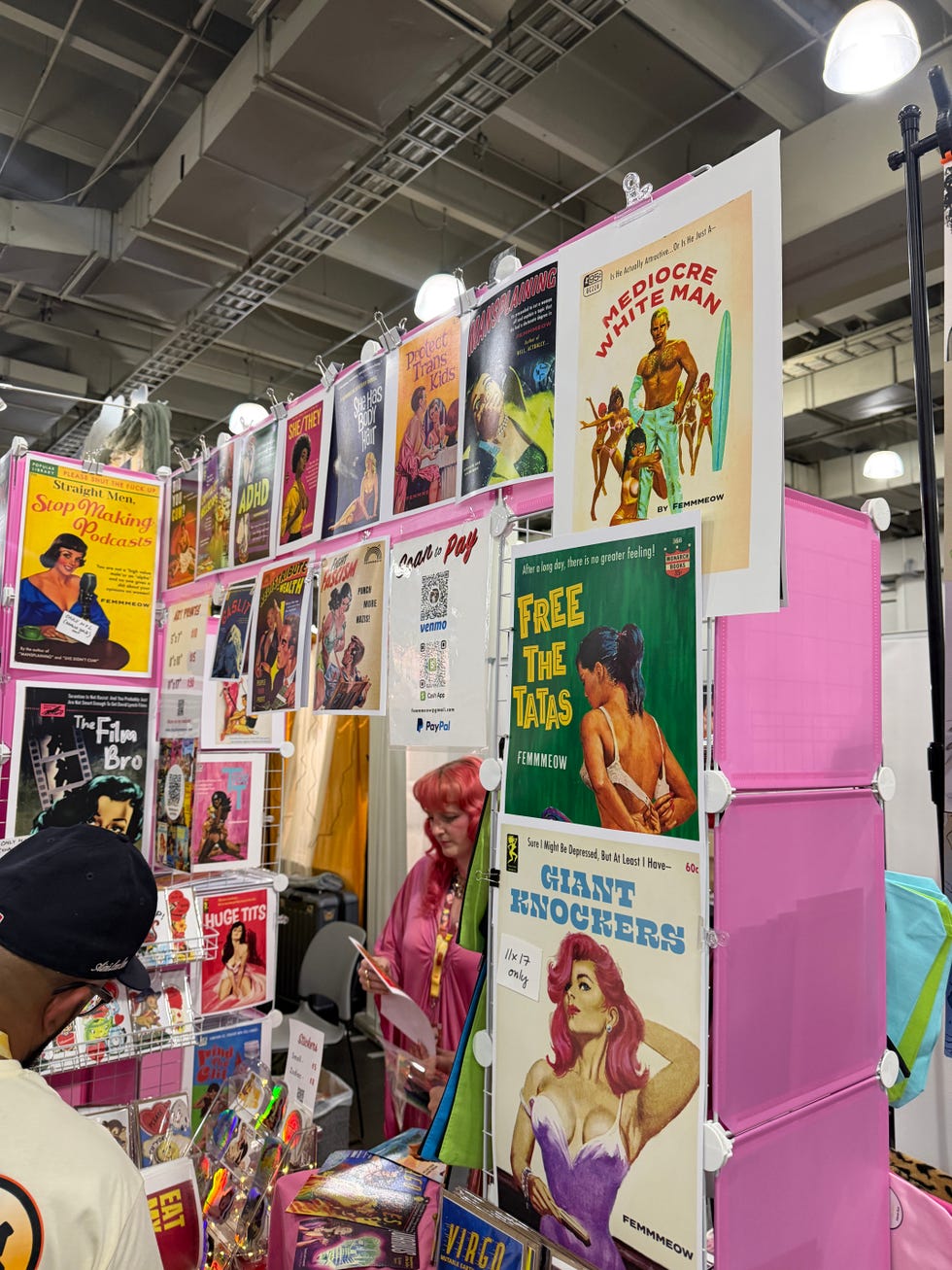 Colorful display of comic books and artwork at an event with a vendor wearing pink hair in the background. Colorful display of comic books and artwork at an event with a vendor wearing pink hair in the background.
