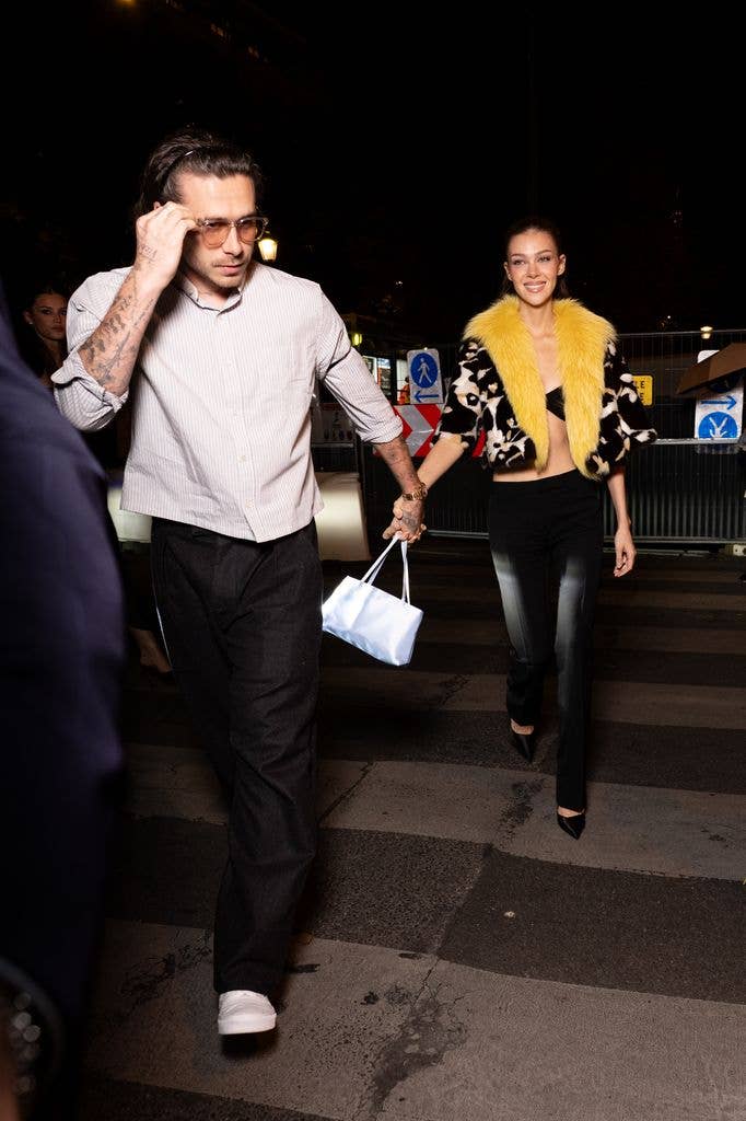 Brooklyn Beckham and Nicola Peltz walking together, he wears glasses and a striped shirt, she has a yellow fur collar.