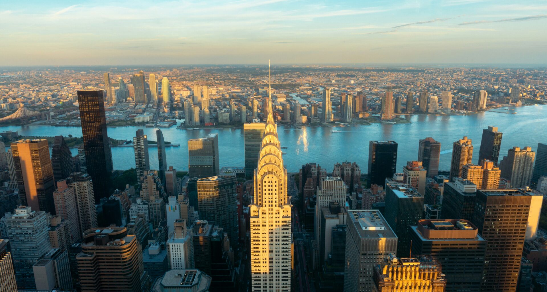 Aerial view of New York City skyline featuring the Chrysler Building, with the East River and Queens in the background under a blue sky at sunset.