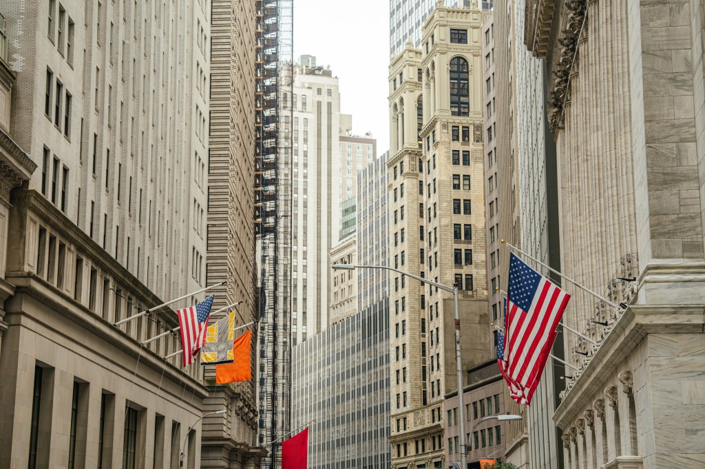 Historic financial buildings and skyscrapers line Wall Street in the New York City financial district, with American flags hanging along the street.