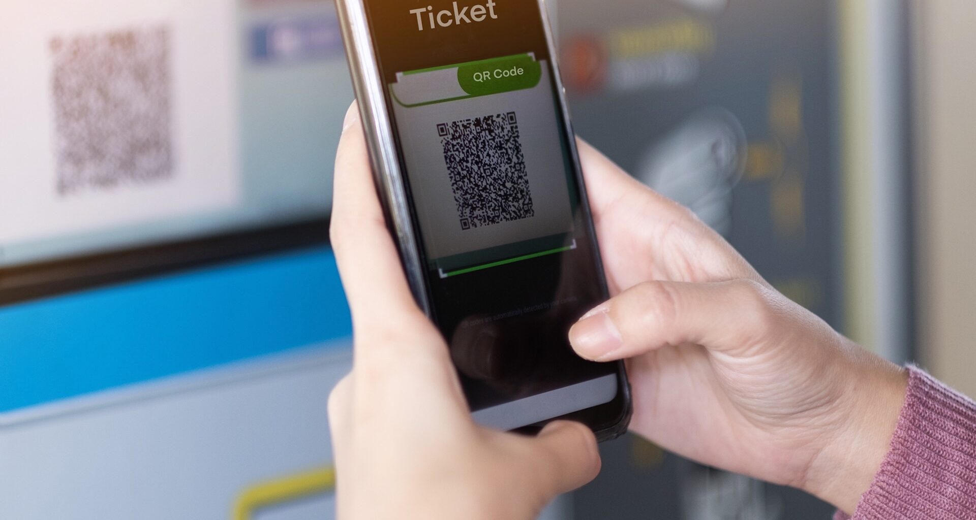 A person scans a QR code on a smartphone in front of a ticket machine.