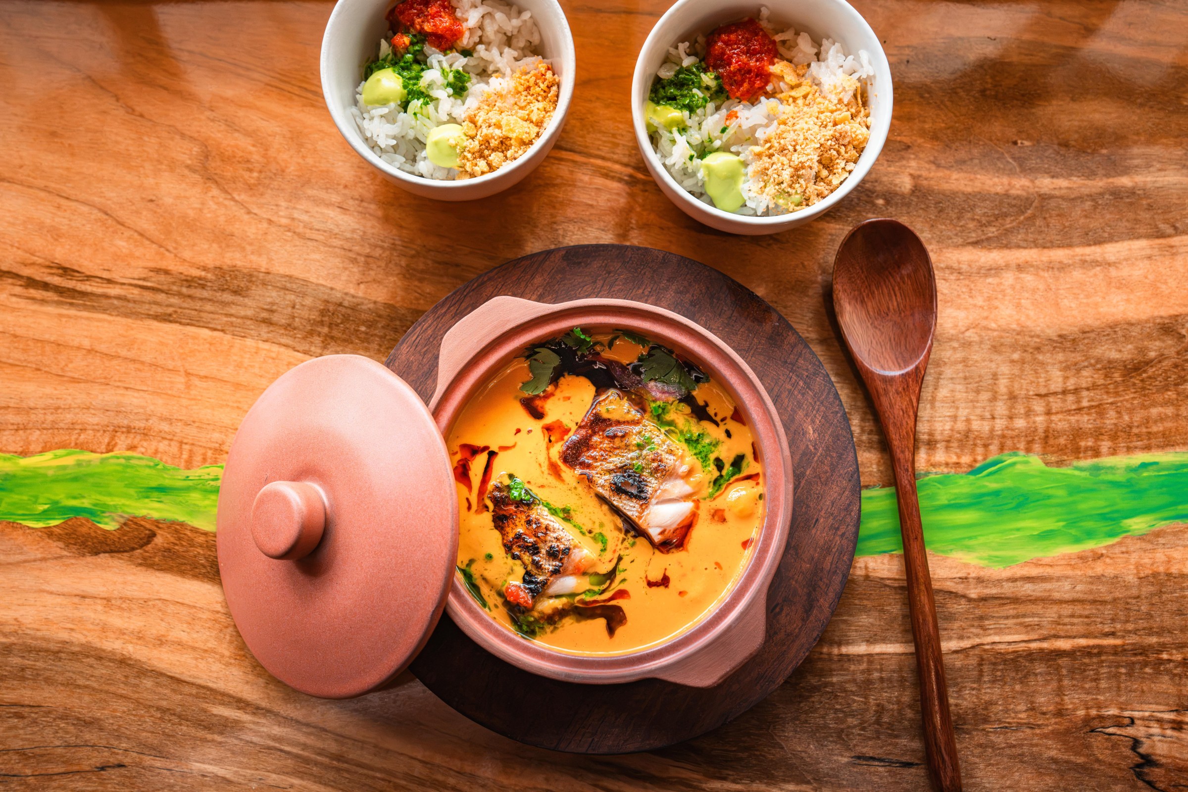 A pot with orange creamy stew with its cover mostly off next to two little bowls of vegetables and rice.