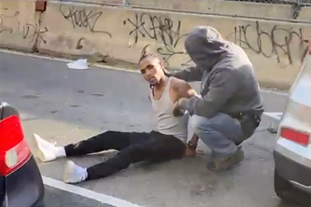 A person in a gray hoodie and jeans kneeling beside and detaining a man sitting on the pavement in a tank top and dark pants next to a graffiti-covered wall.