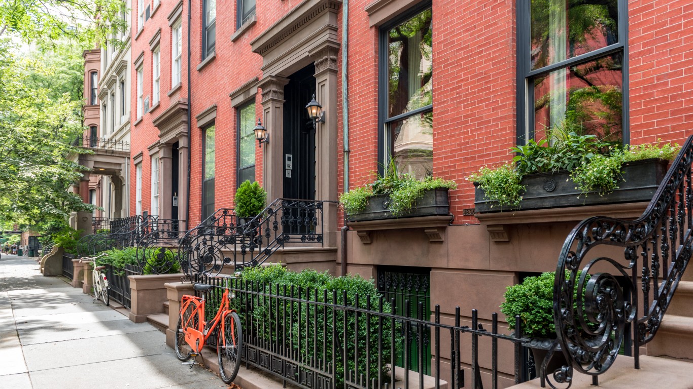 A street view of a row of red brick townhouses with brownstone stoops and ornate black iron railings. Lush green trees line the sidewalk, and planters with greenery adorn the windows. An orange bicycle is parked on the sidewalk in the foreground. The sunlight creates dappled shadows on the pavement.