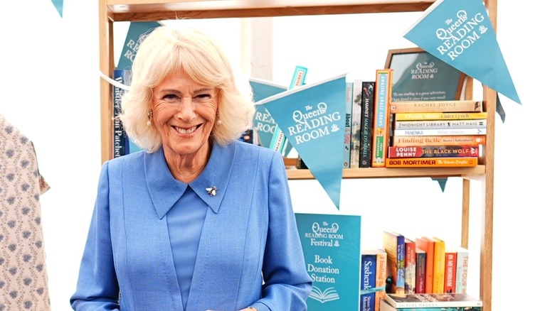 Queen Camilla smiling next to a bookshelf with multiple books at "The Queen's Reading Room" bunting