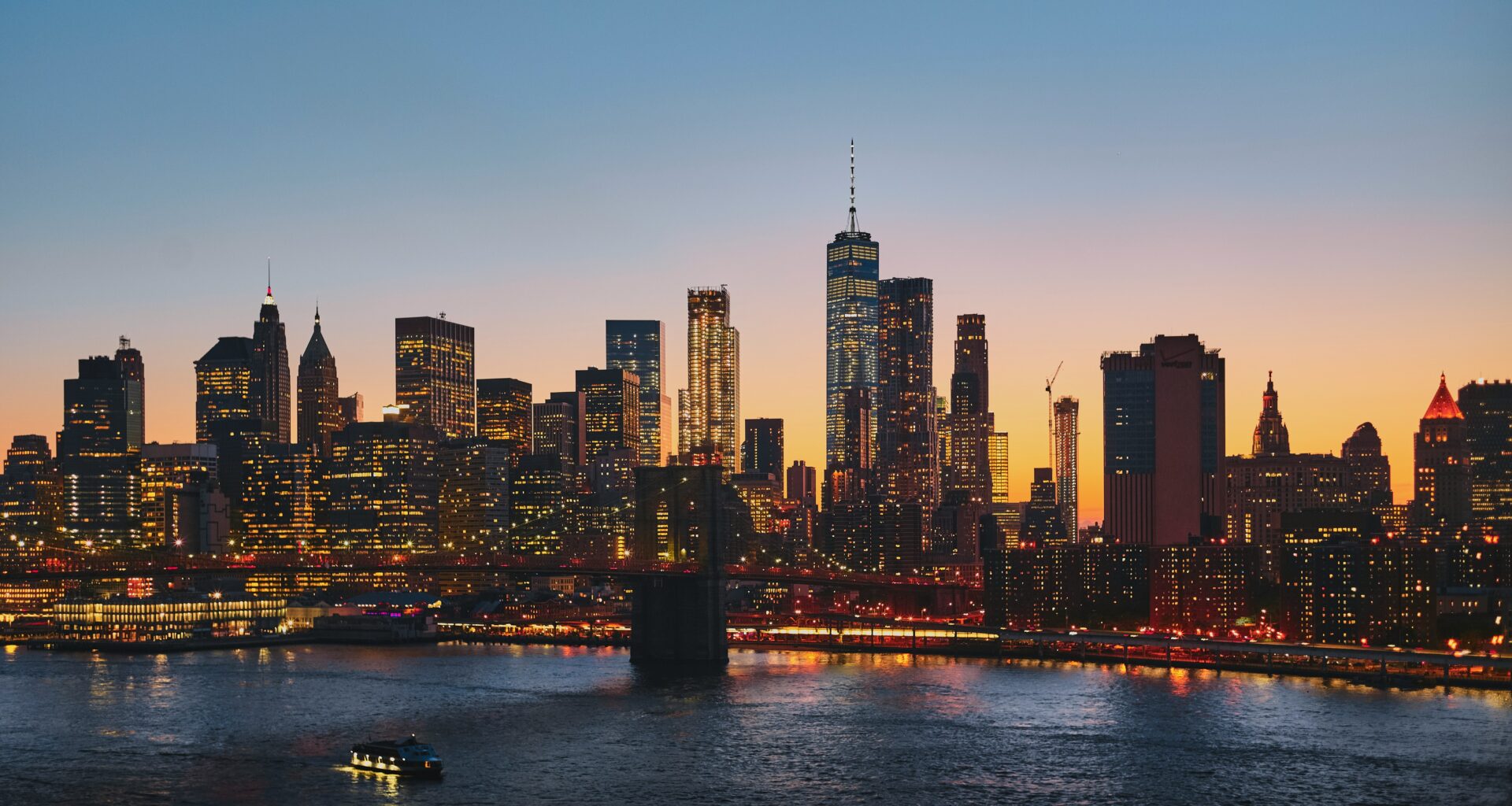 City skyline at sunset with illuminated skyscrapers, a river below reflecting lights, and a small boat in the foreground.
