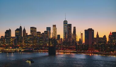 City skyline at sunset with illuminated skyscrapers, a river below reflecting lights, and a small boat in the foreground.