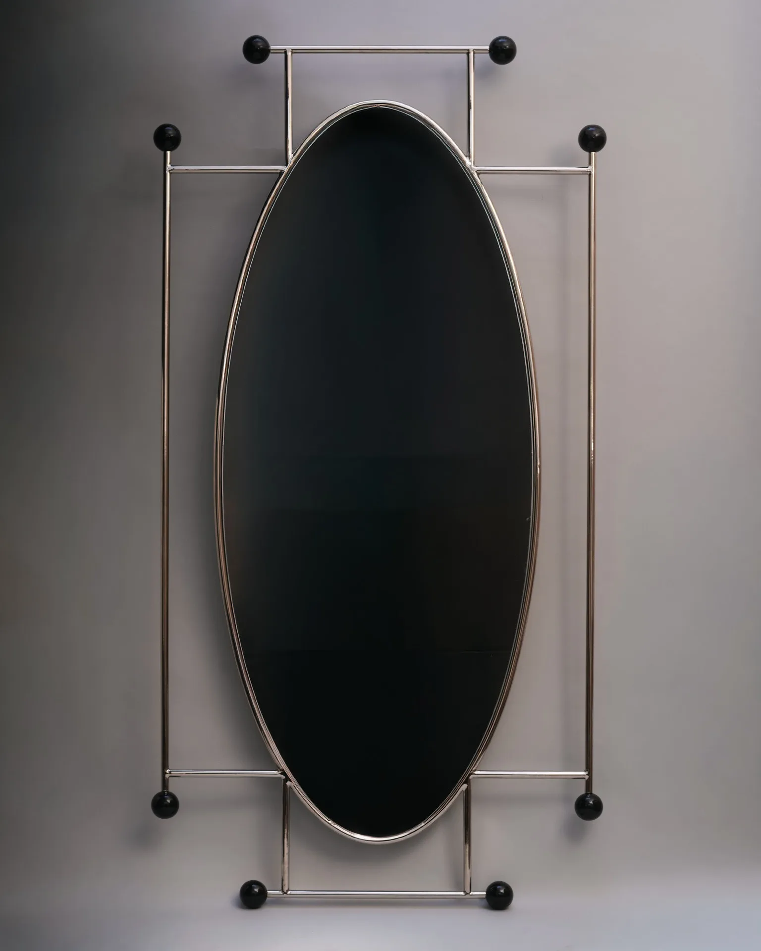 Large oval-shaped mirror with metallic frame and black spherical accents on a gray background.