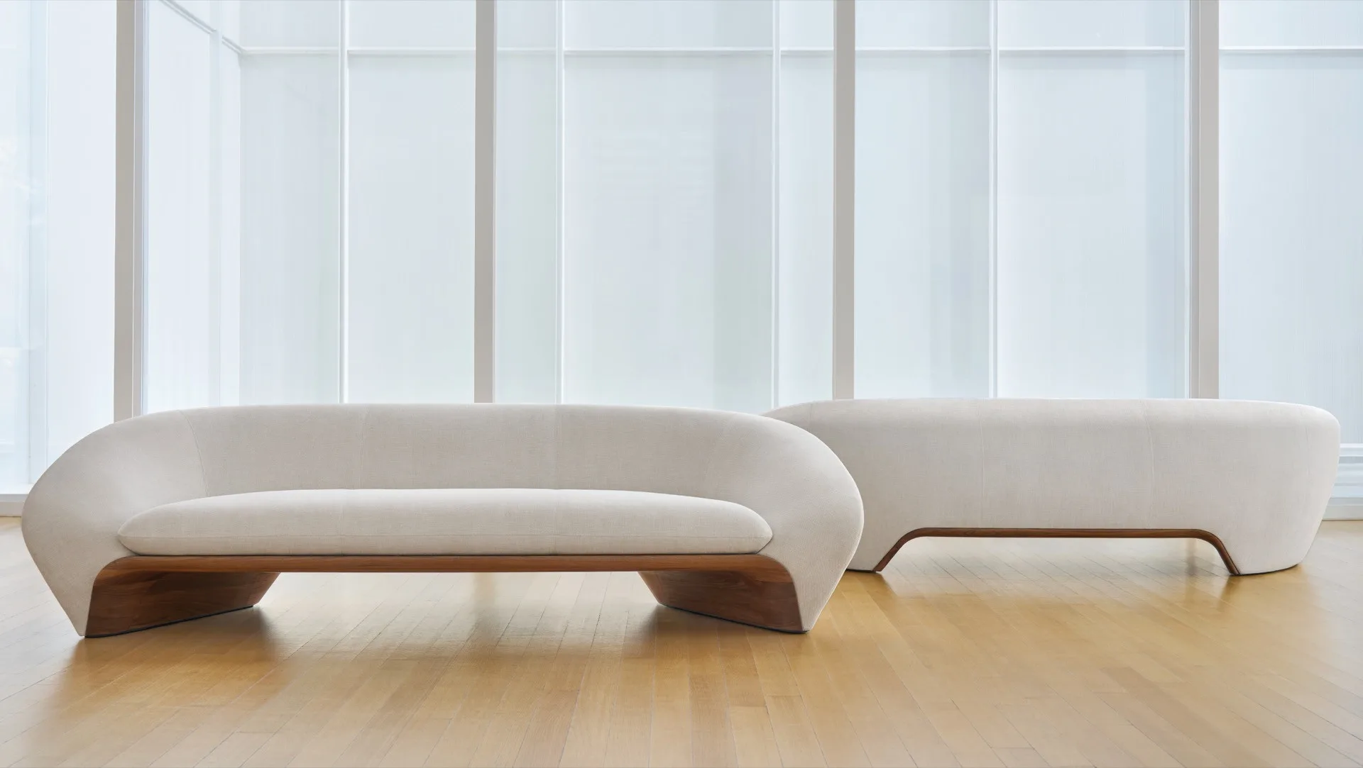 Two curved white sofas with wooden bases on a light wood floor by frosted glass windows.