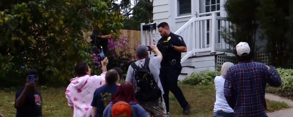 Onlookers watch as a police officer stands outside a home during a standoff.