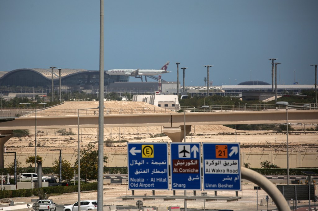 A Qatar Airways plane landing at Hamad International Airport, with road signs in the foreground directing traffic towards Al Corniche, Al Wakra Road, and Old Airport.