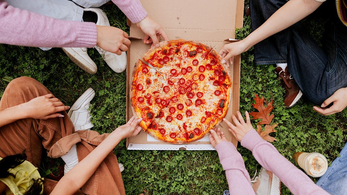 A group of people takes slices of pizza from a box on the grass.