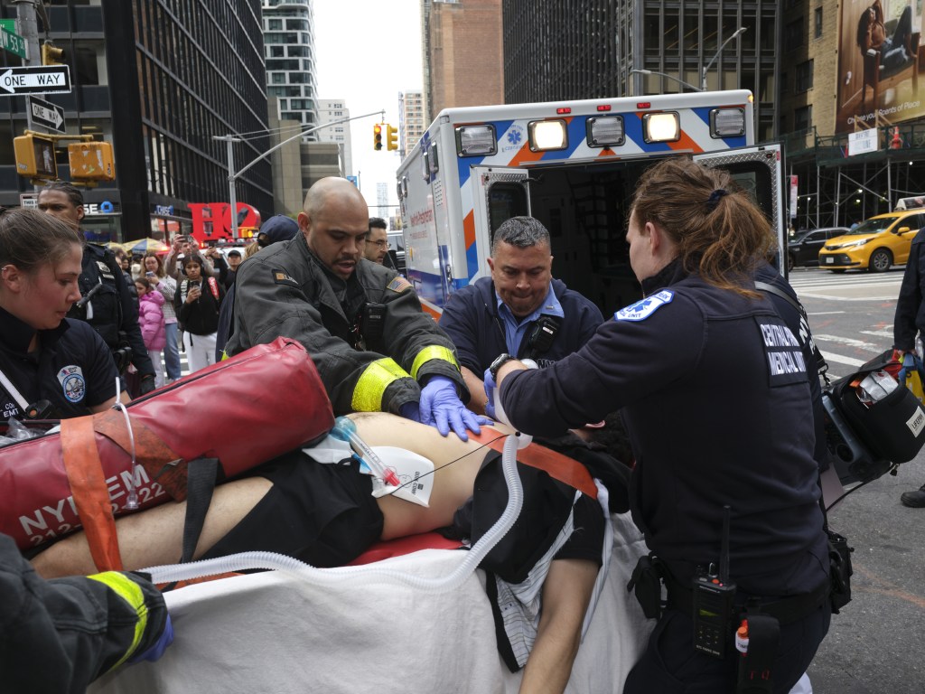 Emergency responders and firefighters tend to a patient on a stretcher in front of an ambulance in a city street.