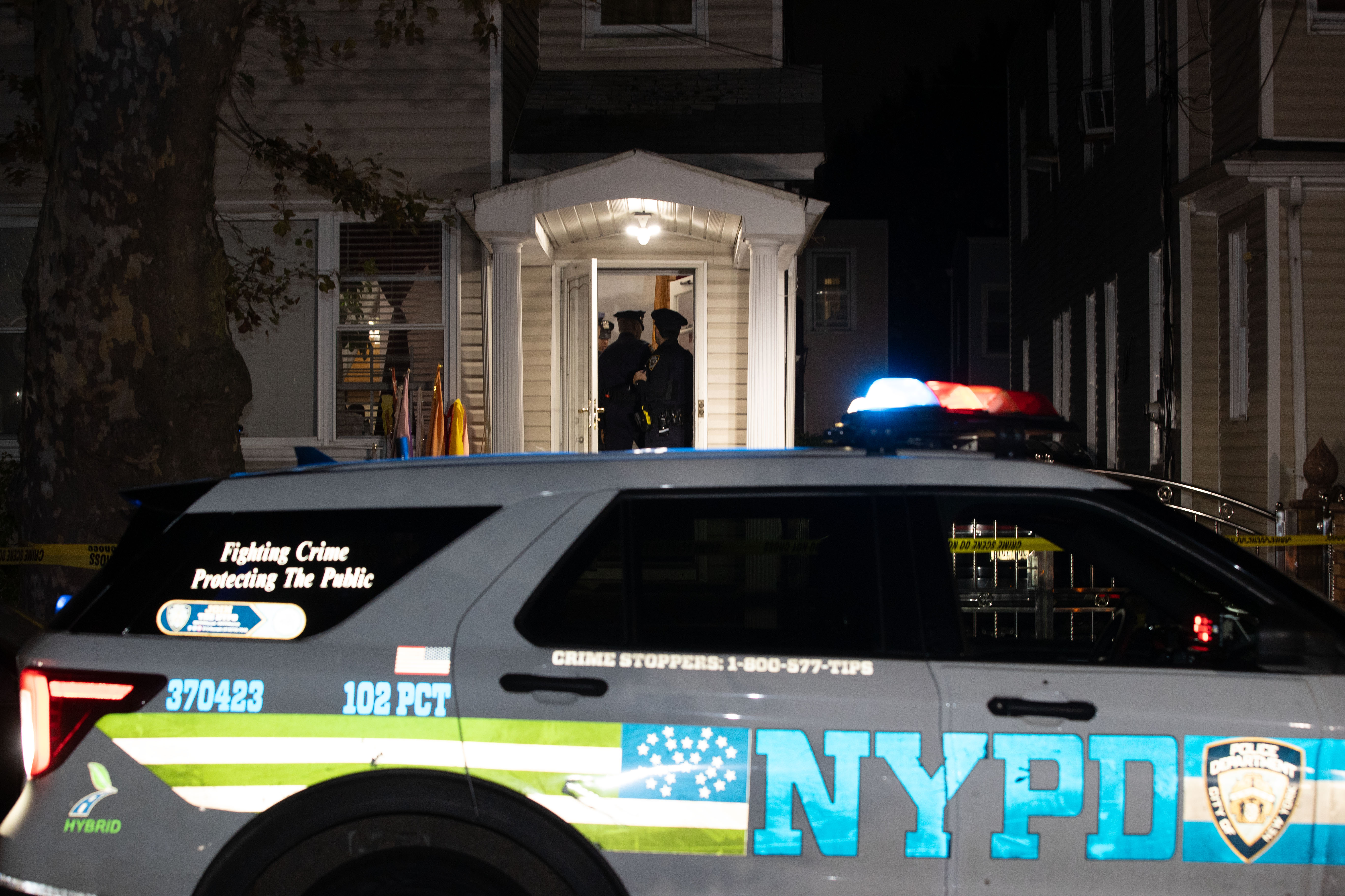 Police officers at the entrance of a home in Queens, with an NYPD patrol vehicle parked in the foreground at night.