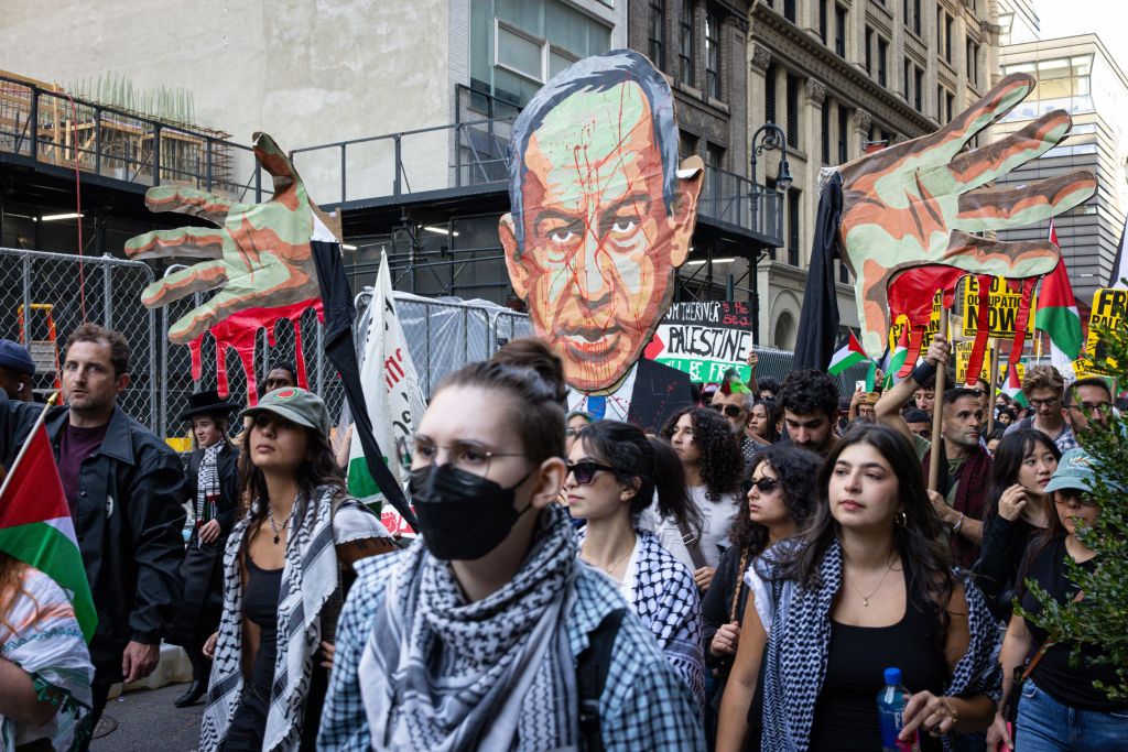 Demonstrators march in a street holding Palestinian flags, large cutouts of hands dripping with red paint, and a caricature of a leader with blood splatters on the face.
