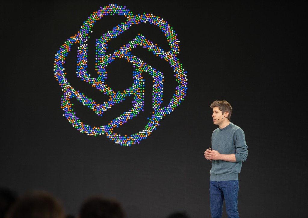 Sam Altman speaks on stage at an OpenAI developer conference in front of a large, colorful abstract logo.