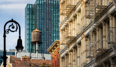 A historic building with external metal fire escapes stands next to a modern Manhattan condo skyscraper, with a rooftop water tower and a vintage street lamp in the foreground.