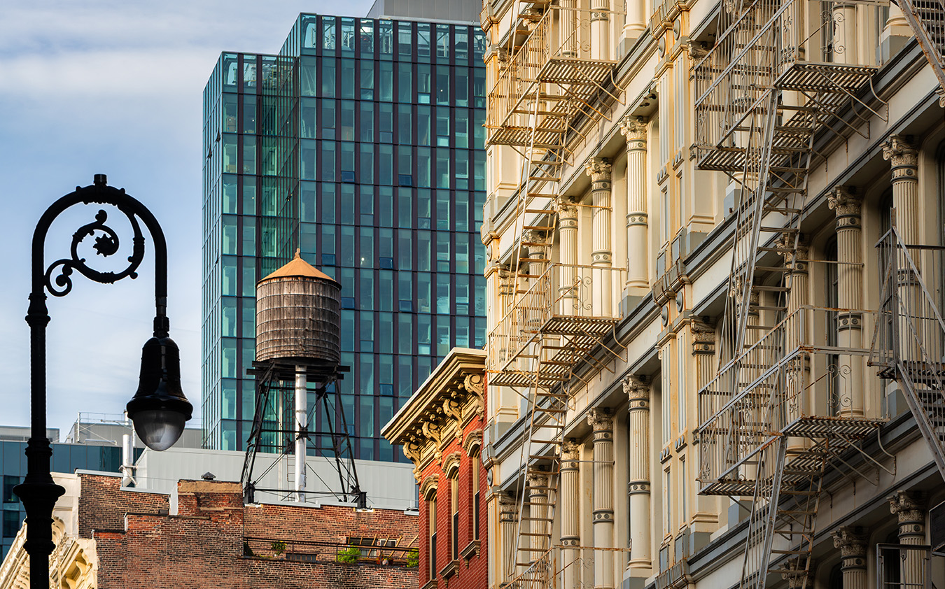 A historic building with external metal fire escapes stands next to a modern Manhattan condo skyscraper, with a rooftop water tower and a vintage street lamp in the foreground.