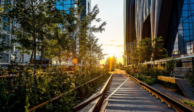 Rail tracks surrounded by plants run between modern glass buildings at sunset, with sunlight streaming through the trees and empty benches on the right, capturing the tranquil ambiance often found near a stylish Manhattan condo.