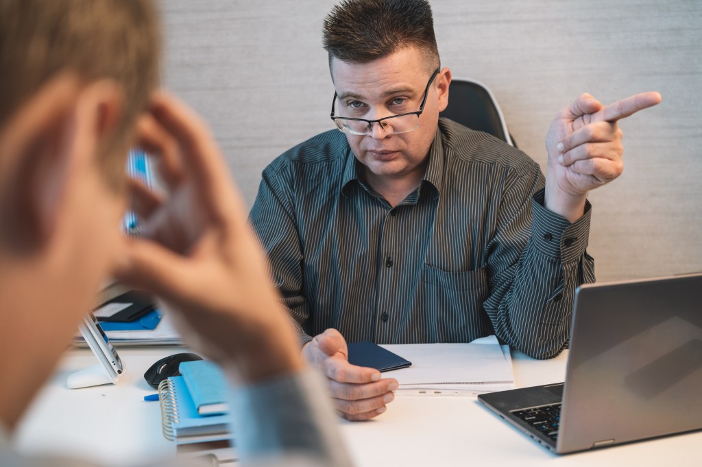 A strict businessman in an office points toward the exit while reprimanding an employee.