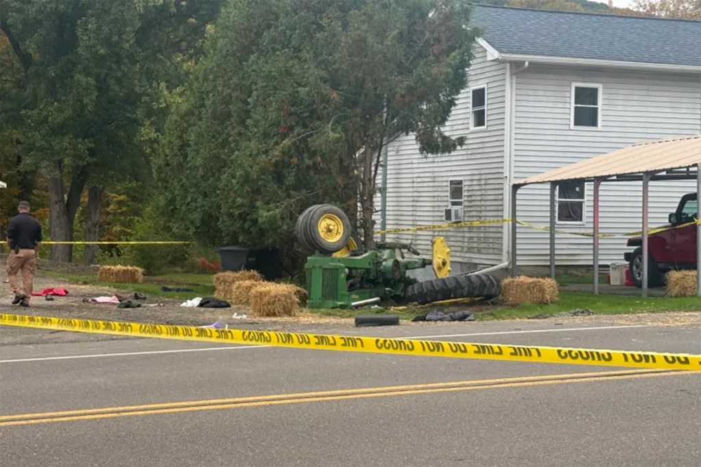 A green and yellow tractor flipped upside down in the front yard of a white house, surrounded by police caution tape.