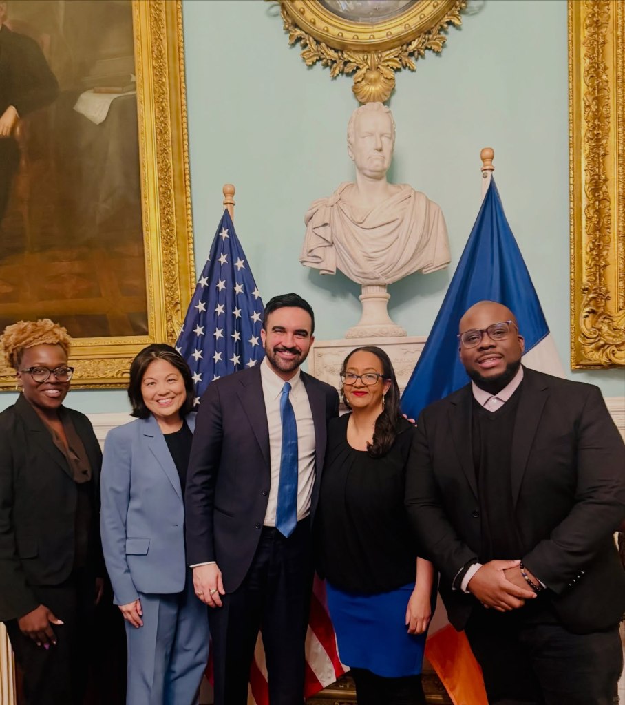 Linda Tigani and Mayor Mamdani pose with three other individuals in a formal room, flanked by American flags and a bust.