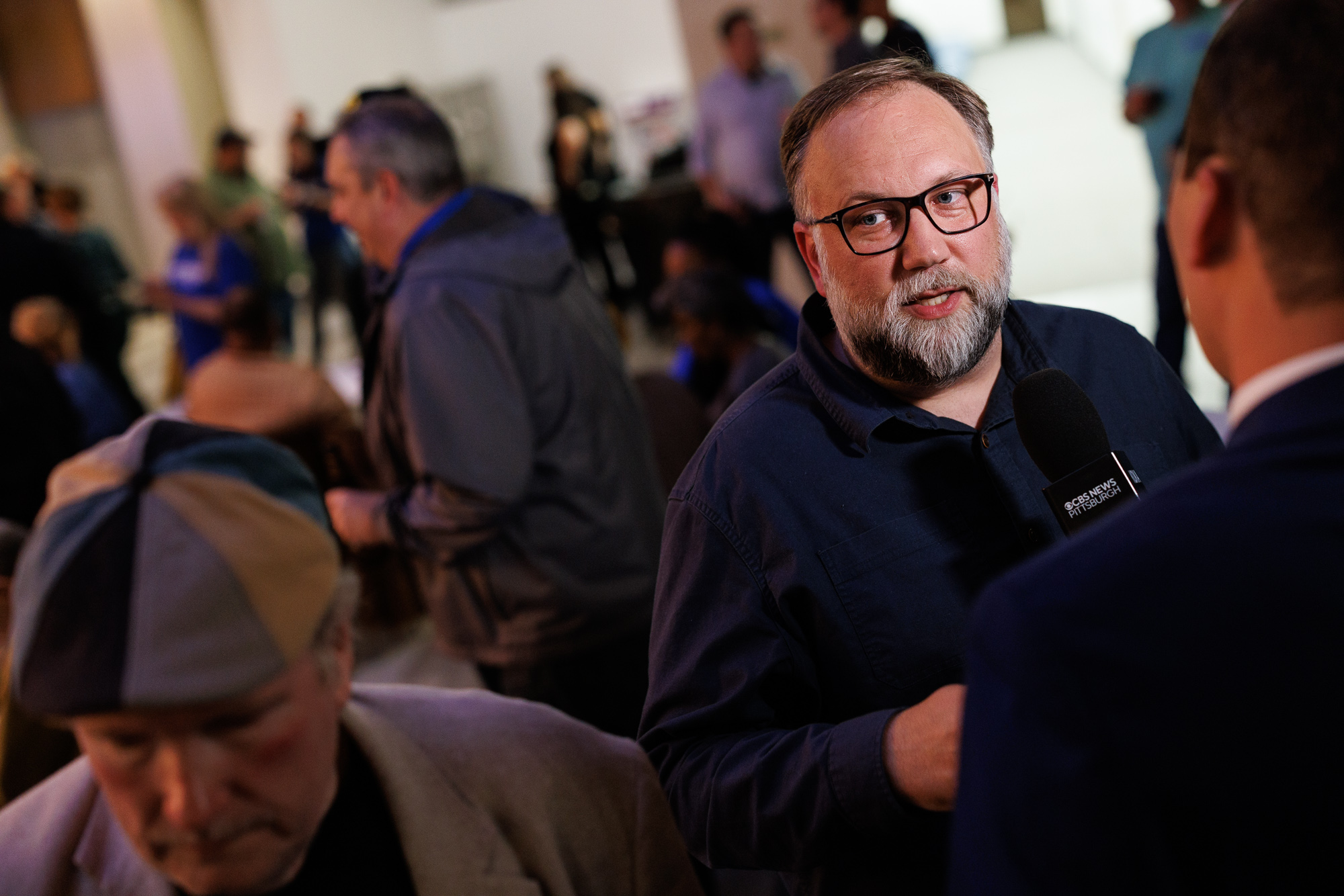 A bearded man in glasses speaks into a microphone during an indoor event, with several people visible in the background.