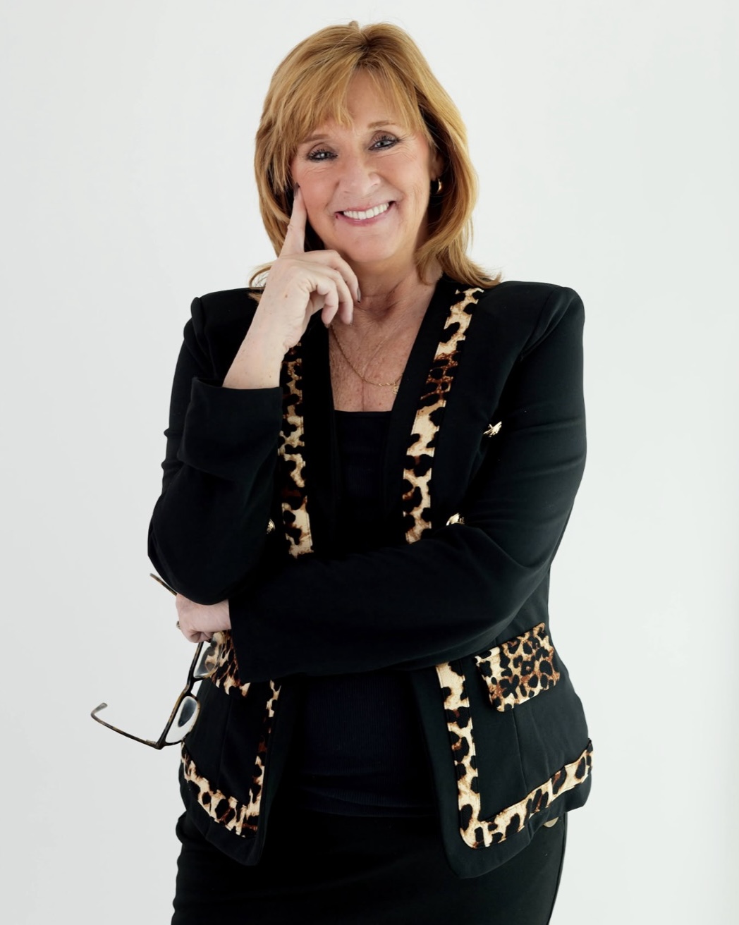 A woman with shoulder-length light brown hair wearing a black blazer with leopard print trim, standing against a white background and smiling, holding eyeglasses in one hand.