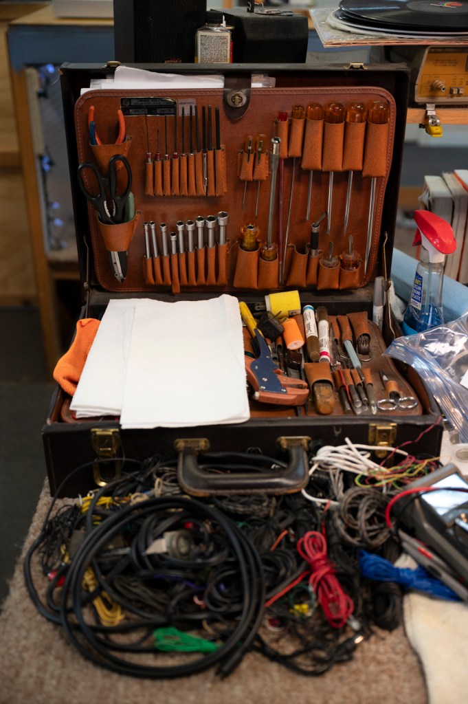 An open, neatly organized leather tool kit on a crowded workbench