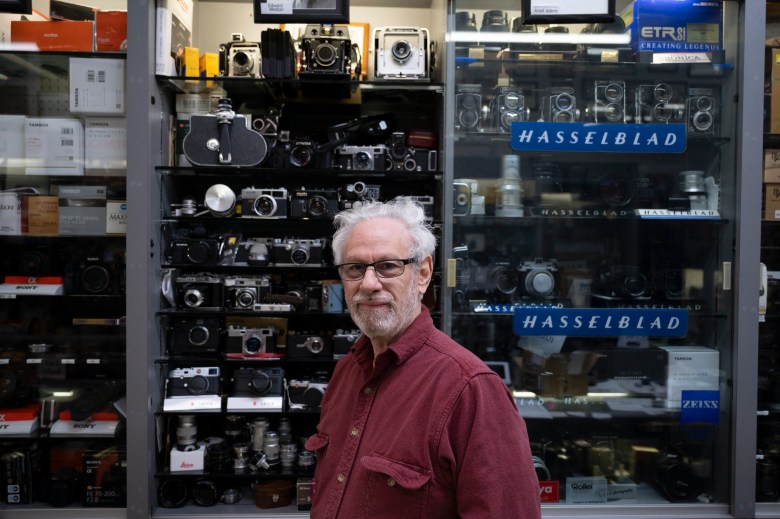 A man with glasses and a beard stands by a glass cabinet full of vintage Hasselblad cameras
