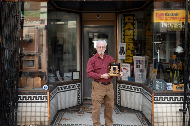 A man with glasses and a beard holds an antique camera outside a Victorian storefront