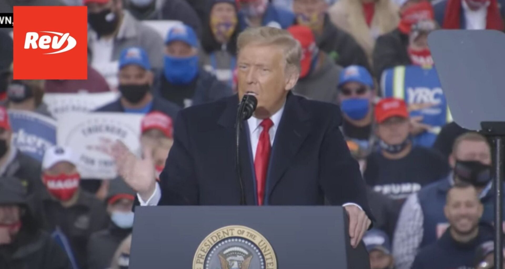 Donald Trump stands and speaks at a podium with a crowd behind him. There is a Rev logo in the top left corner.
