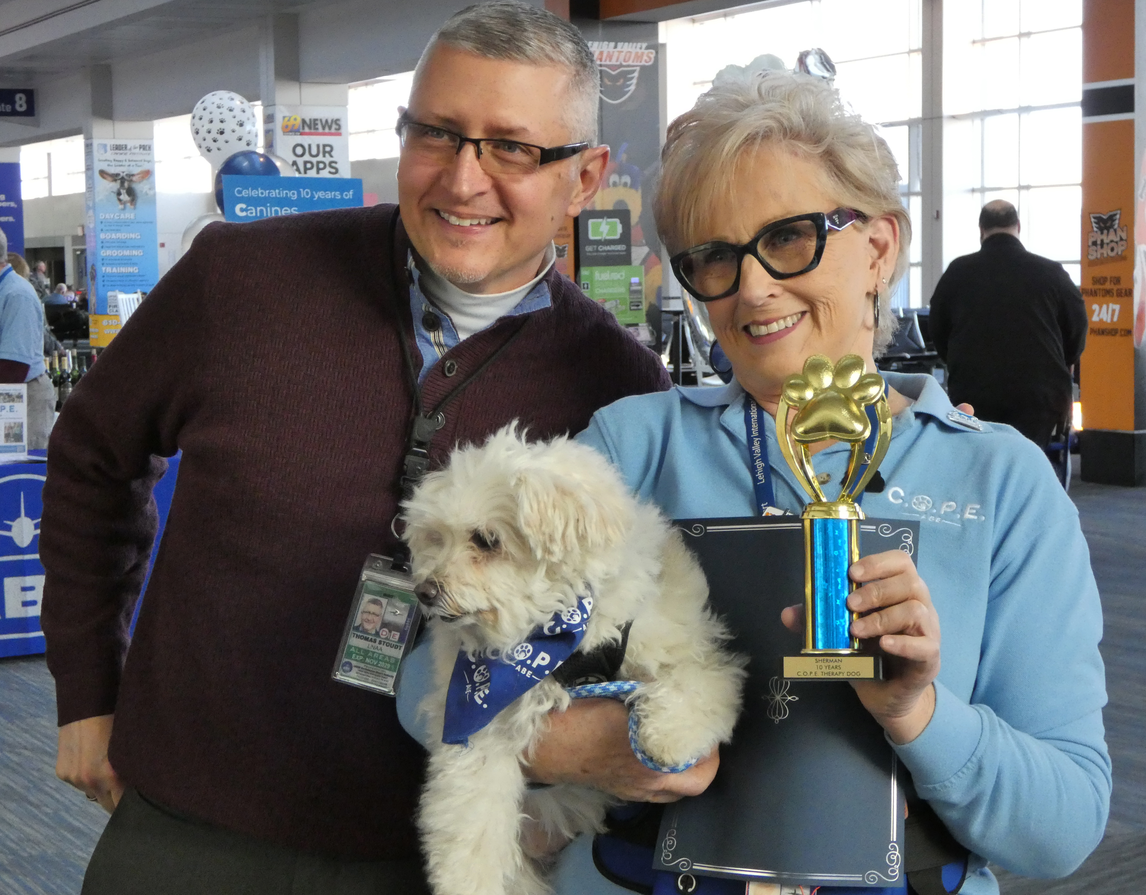 Lehigh-Northampton Airport Authority Executive Director Thomas Stoudt joins Sherman, a Maltipoo (Maltese and the poodle mix), and Sue Ann Reibman during a celebration Thursday, Dec. 18, 2025, of 10 years of Canines Offering Passengers Encouragement (C.O.P.E.) at Lehigh Valley International Airport. Sherman, who is 12, and Reibman have been volunteering with the program since its start in December 2015.