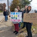 Two women stand along side a road holding protest signs, as more people are gathered behind them.