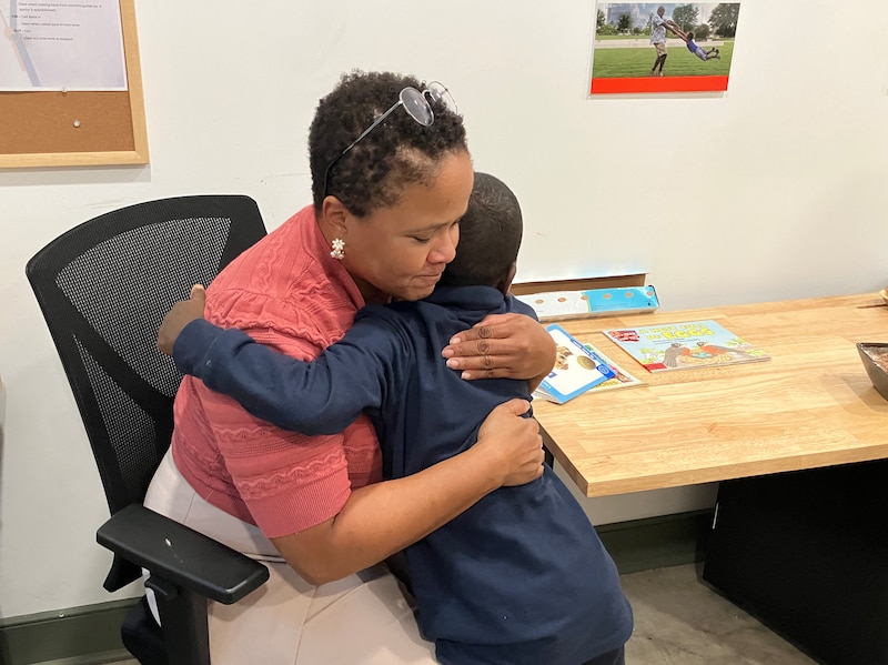 A photograph of a Black woman hugging a young Black boy in a classroom type room.