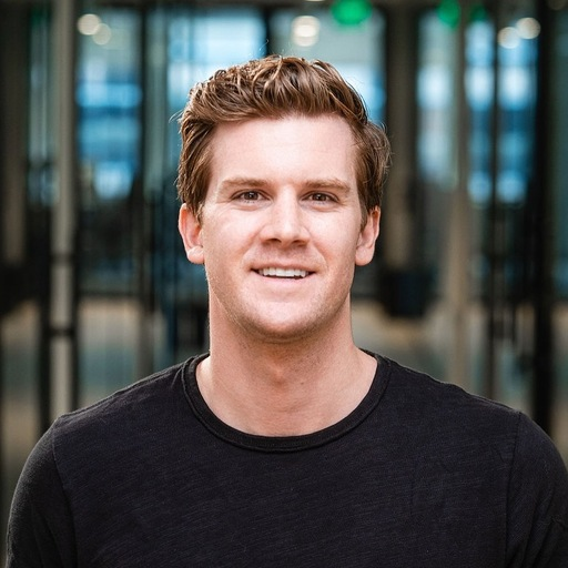 A man with short, light brown hair and a black shirt smiles at the camera while standing indoors with glass walls in the background.