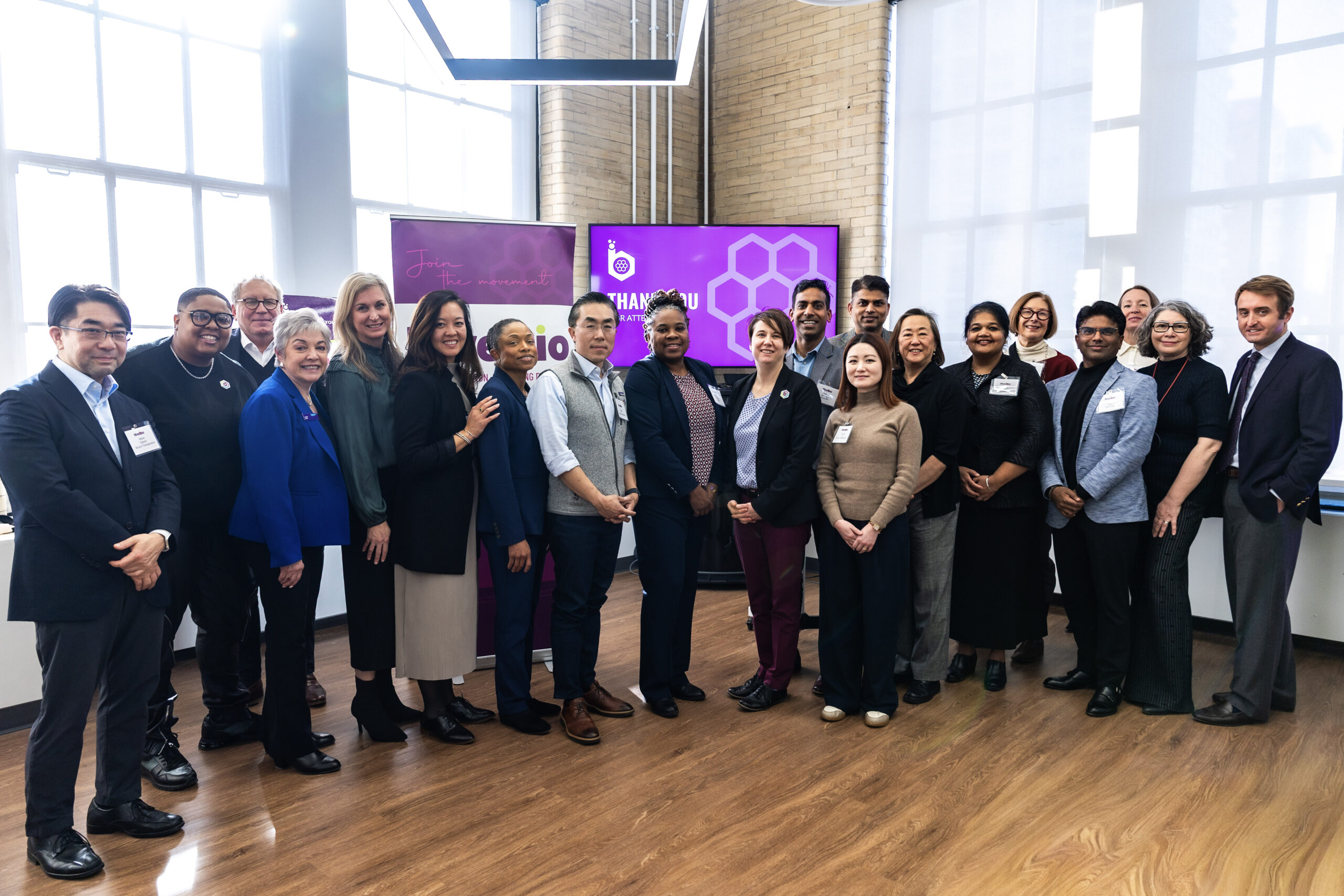 A group of adults, both men and women, stand together in a well-lit room with a wooden floor, posing for a group photo in front of a large screen and windows.