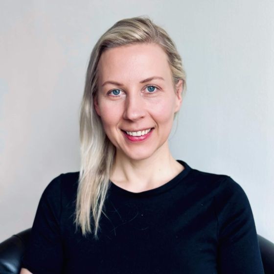 A woman with straight blonde hair and blue eyes, wearing a black top, sits in front of a plain light-colored background, smiling at the camera.