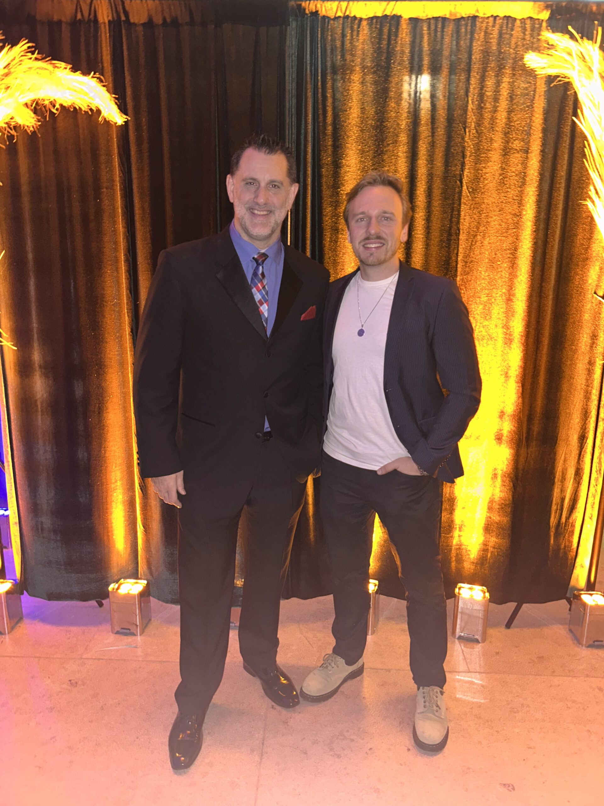 Two men pose together indoors; one wears a black suit and tie, the other sports a white shirt, blazer, and casual shoes. Warm lighting and draped curtains are in the background.