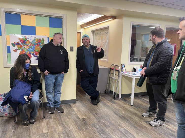 A group of five people are in a room with colorful square decor and a map on the wall. One person speaks animatedly to the others.