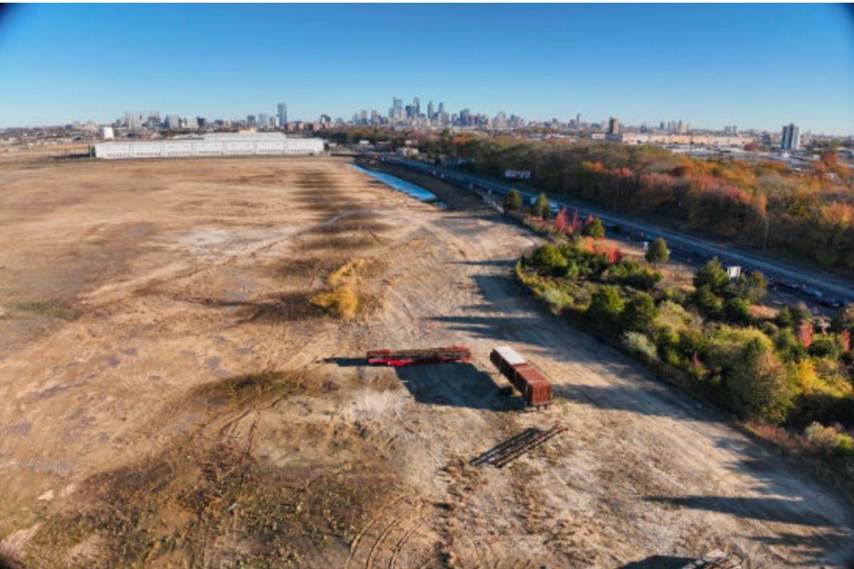 A wide, mostly barren lot with scattered construction equipment and containers, bordered by a road, with a city skyline visible in the distance under a clear sky.