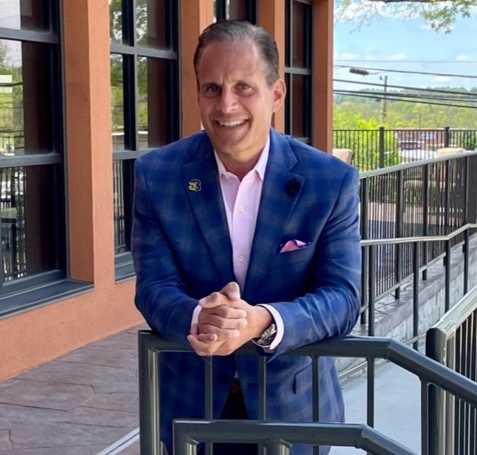 A man in a blue checked suit jacket stands outside by a metal railing, smiling at the camera, with windows and an orange building wall in the background.