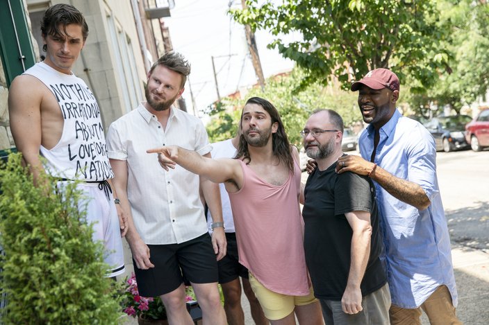 Five men stand next to each other on a tree-lined city street. One in the center, in a pink shirt, points