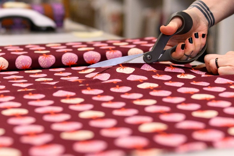 A person with black nail polish uses fabric scissors to cut a piece of maroon cloth with a pattern of pink and white tomatoes.